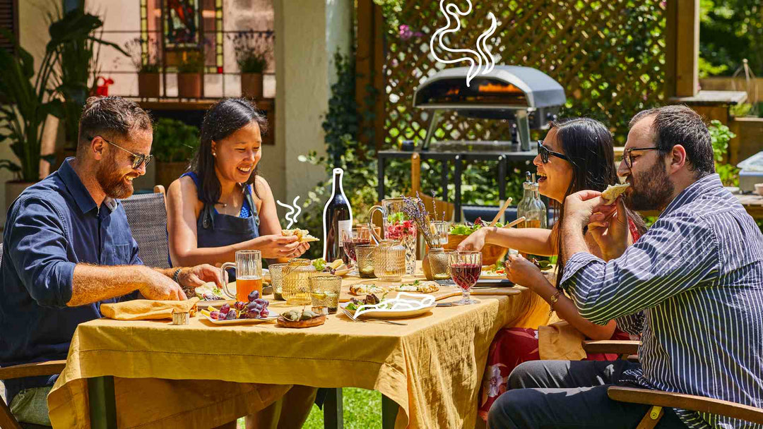 Four people sitting at a table outside eating pizza with an Ooni Koda 16 Gas Powered Pizza Oven in the background.