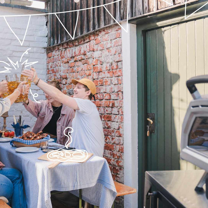 Four people sitting at a table outside and cheering with glasses in the background, a Ooni Karu 16 Multi-Fuel Pizza Oven in the foreground.