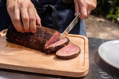 One hand holding the top of a cooked slow-roasted filet mignon, the other hand slicing the steak with a large knife on a wooden cutting board on top of an Ooni Modular Table.