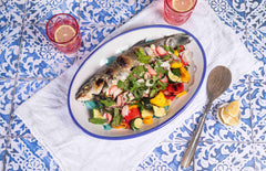 Whole roasted branzino with squash, peppers, shaved spring onions, fennel, radish, parsley, dill and tarragon on a plate on a table.