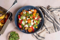 Slow-roasted squash, aubergine, onions, cherry tomatoes and sweet peppers in an Ooni Roasting pan next to a bowl of slow-roasted vegetables with feta and fresh herbs on a table.