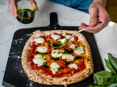 Basil oil being poured out of a glass onto a spoon then drizzled on a pizza topped with tomato sauce and mozzarella