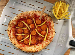 A pizza topped with bratwurst, french fries and German curry sauce on a perforated pizza peel