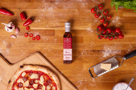 A wooden table top with tomatoes, chillies, garlic, parmesan, chilli oil and pizza showcasing the Ooni groceries range