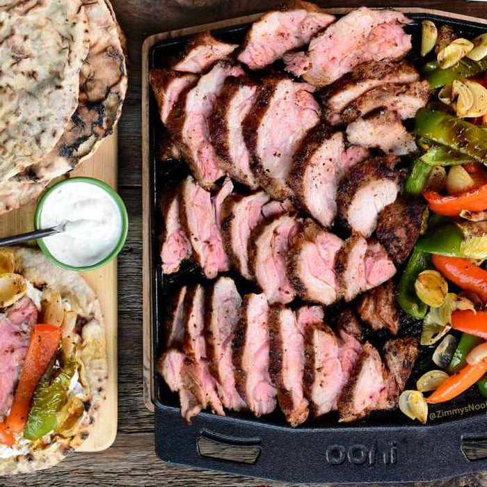 Flatbreads, tahini and harissa spiced lamb neck on a wooden chopping board, next to roasted lamb and vegetables in a cast iron griddle pan. Made using a harissa lamb recipe.