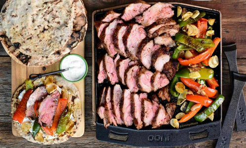 Flatbreads, tahini and harissa spiced lamb neck on a wooden chopping board, next to roasted lamb and vegetables in a cast iron griddle pan. Made using a harissa lamb recipe.