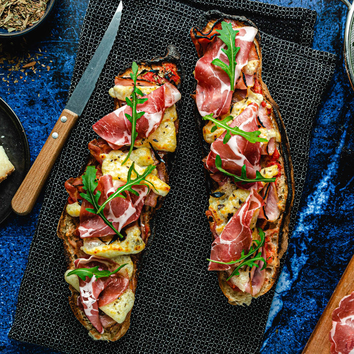 Cooked Corsican pizza baguette with Corsican cheese, pancetta, coppa, rocket and herbes de Provence on a table surrounded by a plate of cheese, bowl of rocket and serving board of pancetta