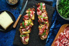 Cooked Corsican pizza baguette with Corsican cheese, pancetta, coppa, rocket and herbes de Provence on a table surrounded by a plate of cheese, bowl of rocket and serving board of pancetta