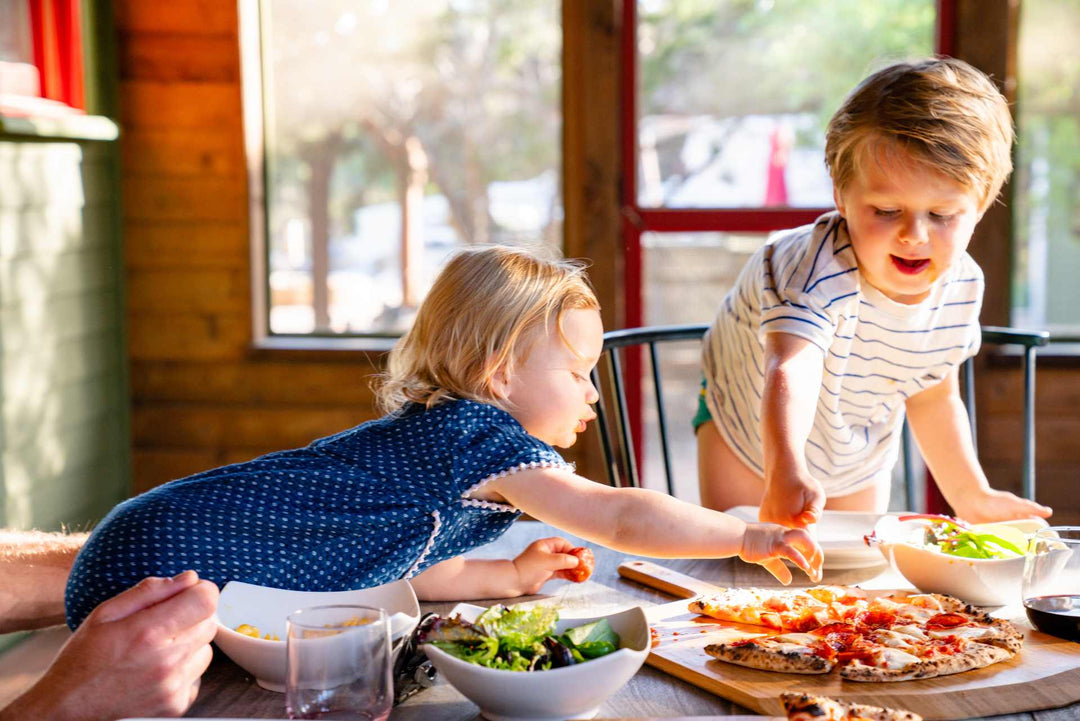 Two young children reaching over a table with a cooked pepperoni pizza on an Ooni Bamboo Peel & Serving Board.