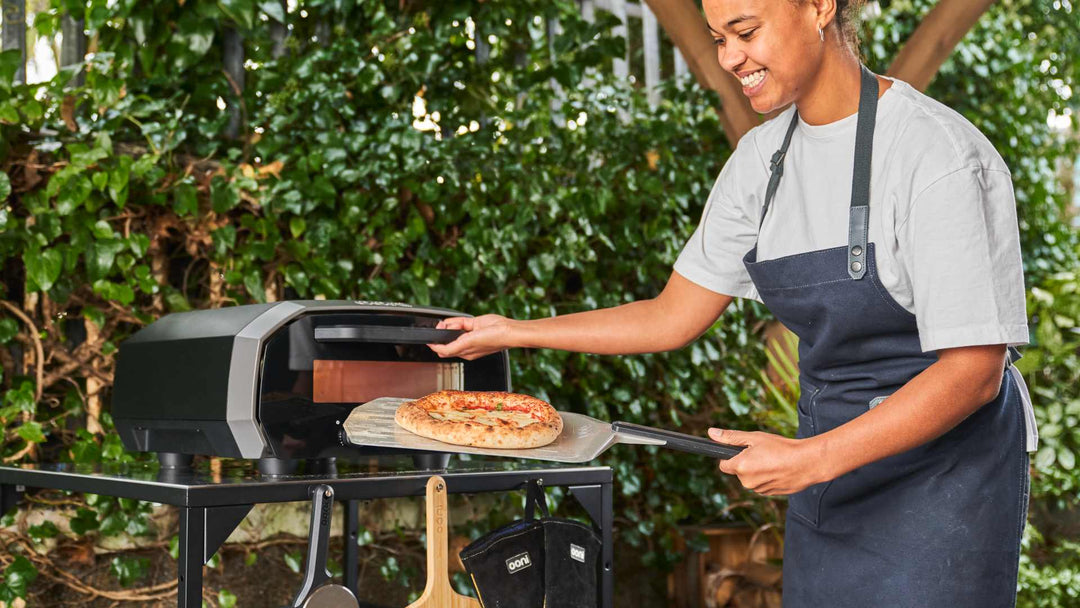 One person holding onto the handle of an Ooni Perforated Pizza Peel with a cooked cheese pizza on top, the other hand holding onto the door handle of an Ooni Volt 12.