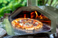 A pizza with topped with steak, blue cheese and red onion on a metal pizza peel being inserted into an Ooni pizza oven. Made using a steak pizza recipe.