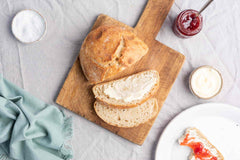 Half of a fresh-baked, no-knead bread loaf and two slices, one spread with butter, all on a bamboo serving board