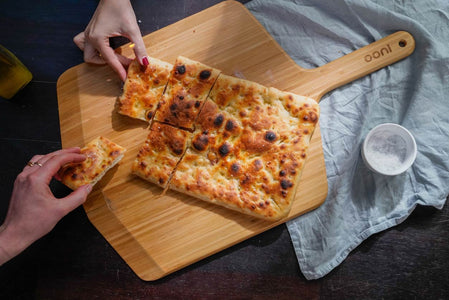 Two hands holding pieces of Roman pizza on an Ooni Bamboo Pizza Peel & Serving Board.