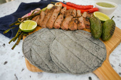 Masienda blue corn tortillas on an Ooni Bamboo Pizza Peel & Serving Board next to green and red chillies, sliced carne asada, grilled onions, sliced limes and salsa verde.